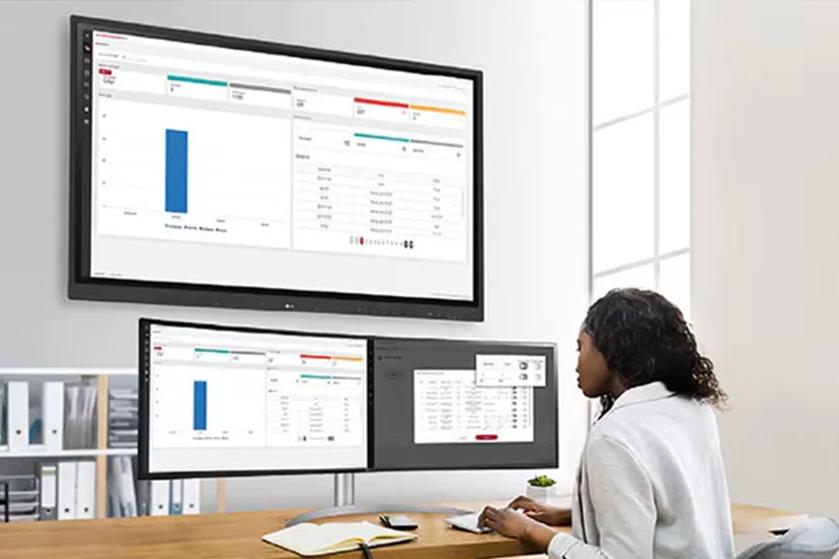 An office worker looks at her monitor. ConnectedCare's UI is displayed on the monitor and the CreateBoard on the wall above her.