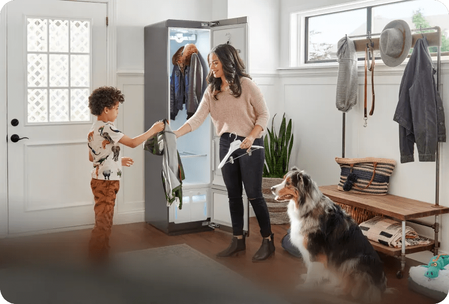 A young boy hands his jacket to his mother who is placing it on a hanger to store in the LG Styler®. A dog sits nearby.