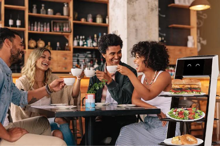 A group of friends sit at a table in a cafe, raising coffee cups in cheers. CLOi ServeBot stands beside the table, delivering various cafe foods. 