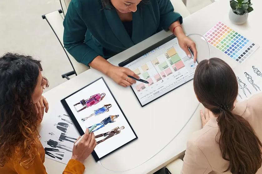 Three people discussing fashion designs using tablets, with sketches, color palette, and a plant on the table.