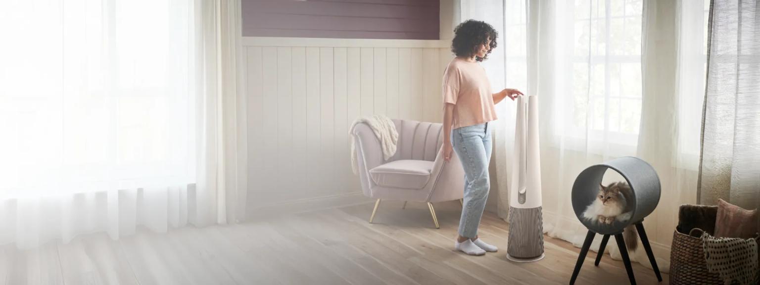 A woman adjusts her PuriCare™ air purifier in a sun-filled room. 