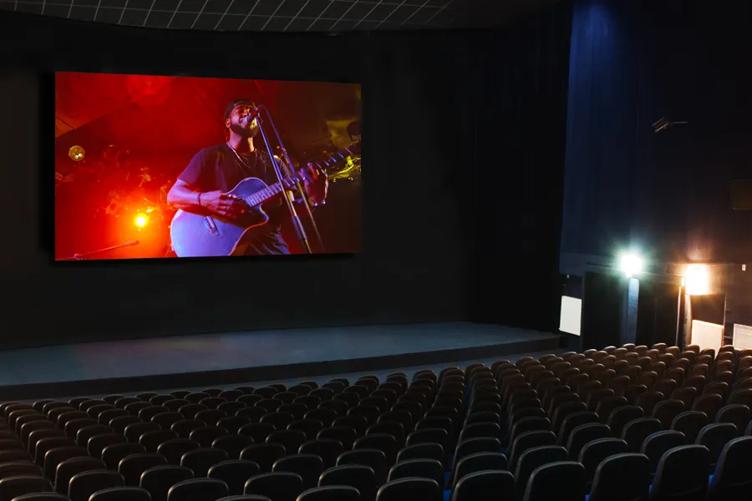 Movie theater showing a performer playing guitar and singing on screen, with rows of empty seats.