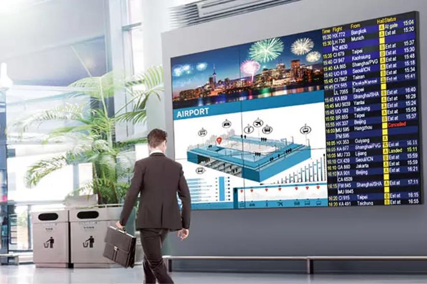 A large digital display board in an airport showing a city skyline, airport map, and flight information. A man with a briefcase walks by.