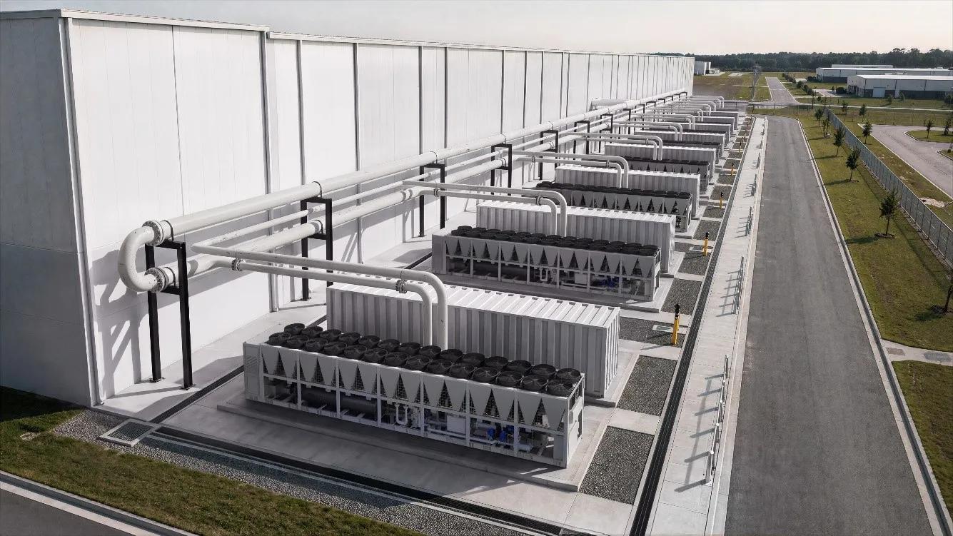 Aerial view of rows of large industrial air-cooled chillers with interconnected piping installed alongside a large commercial facility, set in a flat industrial campus with access roads and green surroundings.