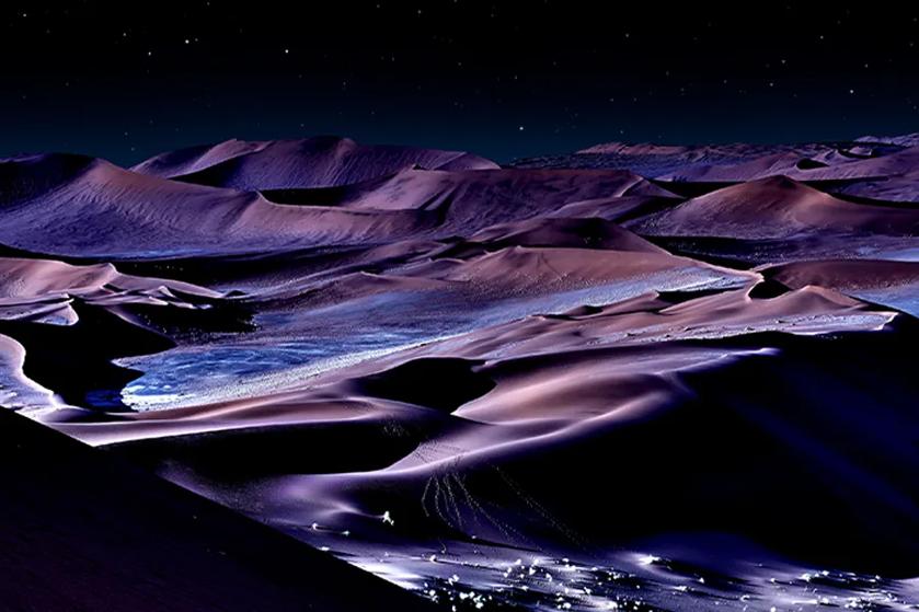 Desert dunes under a starry night sky with purple and blue hues.
