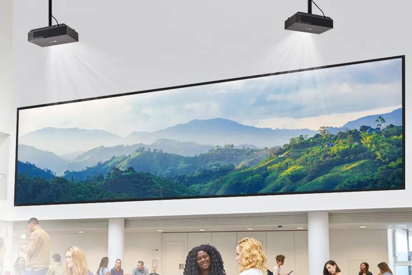 The image shows a bright and modern interior space with a large, panoramic screen mounted on the wall. The screen displays a vivid landscape of rolling green hills and distant mountains under a partly cloudy sky. Two ceiling-mounted projectors are positioned above, casting light toward the screen. Below the screen, several people are engaged in conversation in a well-lit room with white walls and a white ceiling. The room has wide rectangular vents near the ceiling.