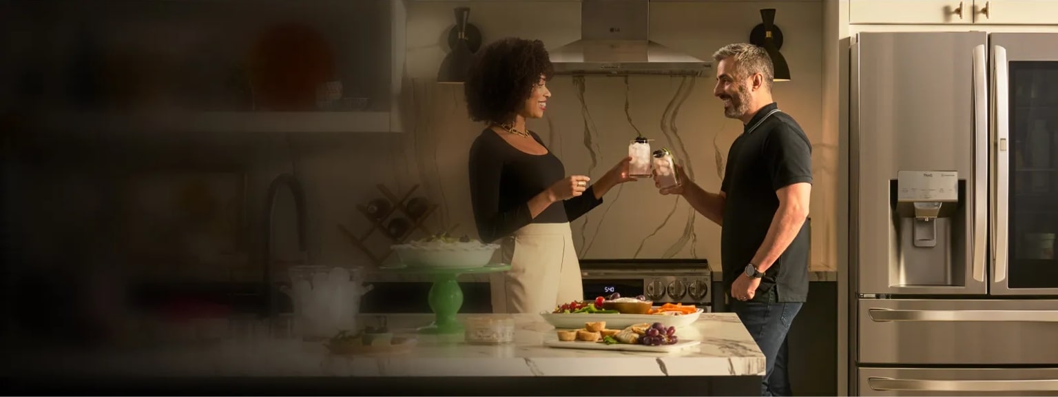 A couple "cheers" their drinks in front of their LG refrigerator in a modern kitchen.