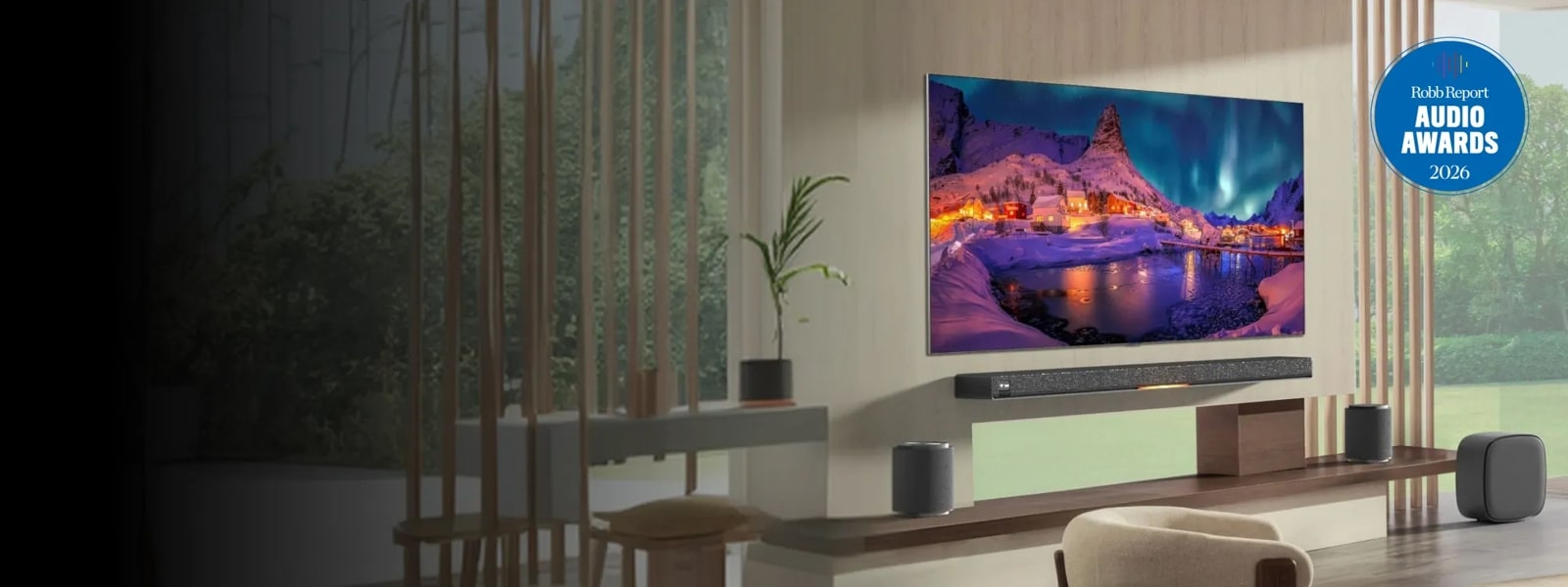 LG TV mounted on a wall displaying a snowy northern lights scene, with an LG Soundbar on a credenza below and multiple LG speakers arranged around it in a modern living room setting, with Robb Report Audio Awards 2026 badge visible.