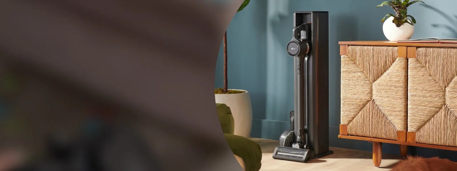 Alt text: A sleek LG CordZero cordless vacuum in a vertical charging stand placed next to a woven-front wooden cabinet and indoor plants in a modern living room.