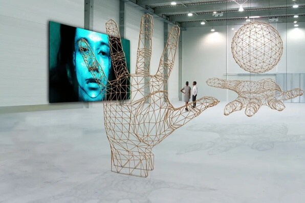 A spacious room with wireframe sculptures of hands and a sphere, and a digital screen showing a blue-toned face.