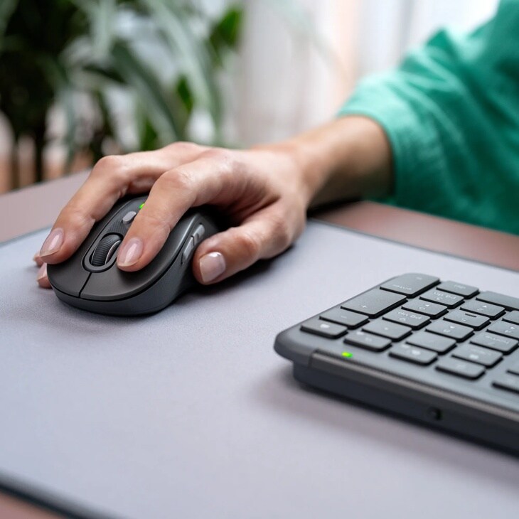 Logitech 920013271 Hand using wireless black mouse on gray mousepad next to keyboard with green indicator light, person wearing teal sleeve
