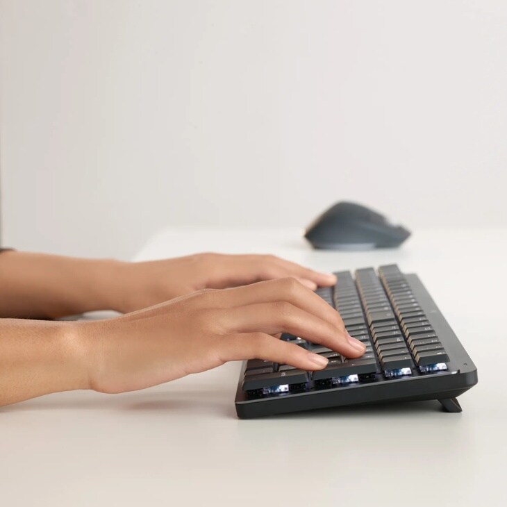 Hands typing on 920010549 wireless keyboard with backlit keys and wireless mouse visible in background on white desk