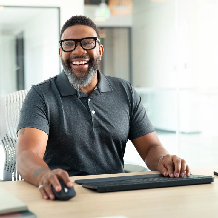Person in gray polo shirt smiling while using wireless keyboard and mouse at desk in modern office setting - 920010923
