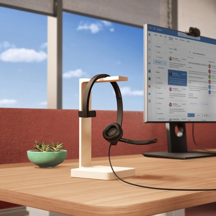 Logitech 981000570 headset with microphone on white stand next to monitor displaying email interface in bright office workspace