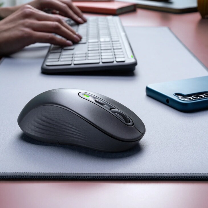 Logitech 920013271 wireless mouse with textured surface and green indicator light on desk with keyboard and smartphone