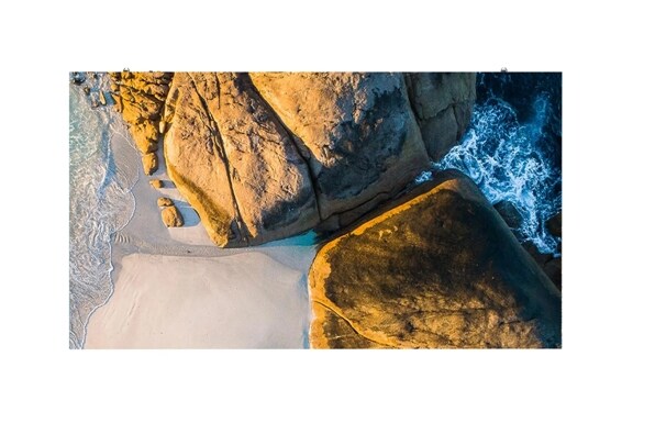 Aerial view of golden-brown coastal rocks, pale sand, and foaming blue ocean waves.