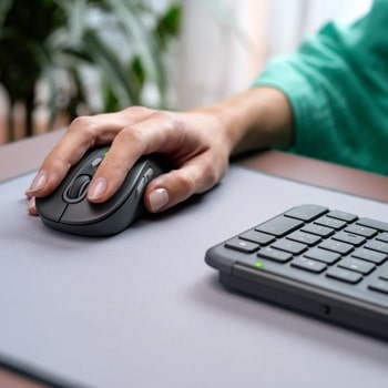 Logitech 920013271 Hand using wireless black mouse on gray mousepad next to keyboard with green indicator light, person wearing teal sleeve
