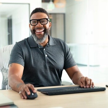 Person in gray polo shirt smiling while using wireless keyboard and mouse at desk in modern office setting - 920010923