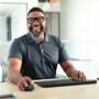 Person in gray polo shirt smiling while using wireless keyboard and mouse at desk in modern office setting - 920010923