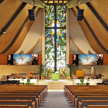 Interior of a modern church with stained glass, organ pipes, and screens displaying images of singers and a dove.1