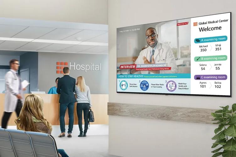 A woman sits in a hospital waiting area, looking at an LG Hospital TV. It displays an interview with a doctor, wayfinding information, and tips on staying healthy. 