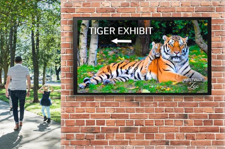 An external brick wall takes up most of the image. On it is an LG outdoor display, with a photograph of a tiger and a cub, an arrow, and the words 'Tiger Exhibit.' Beyond the wall, a father and child are walking on the zoo's path. 