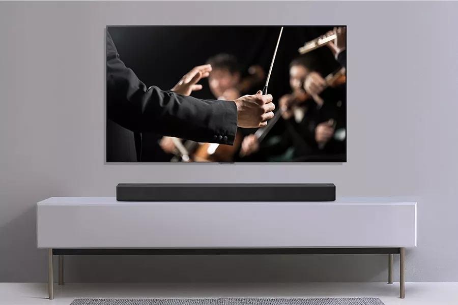 A TV is shown on a gray wall and LG Soundbar below it on a gray shelf. TV shows a conductor conducting an orchestra.