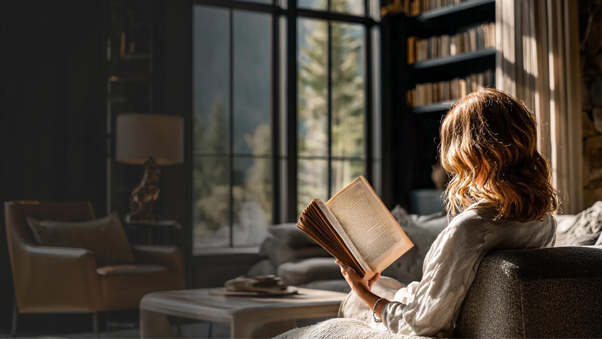 Person relaxing on a sofa reading a book in a sunlit living room with large windows and bookshelves