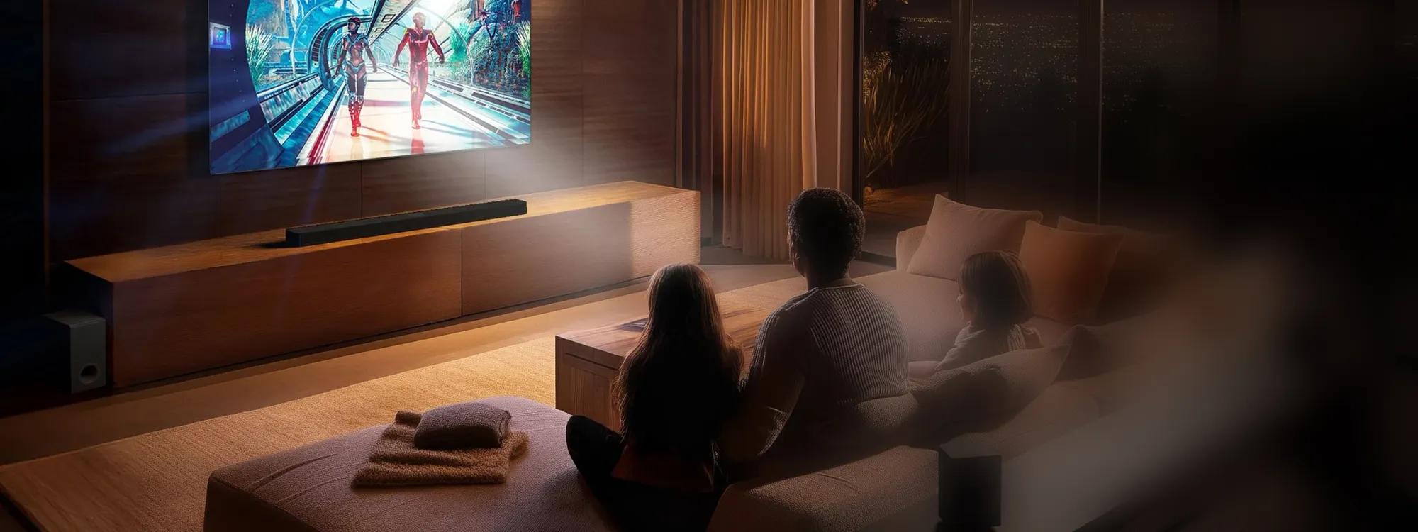 Father and daughter sitting on a sofa in a darkened living room watching a sci-fi scene on large wall-mounted TV with a soundbar and subwoofer.