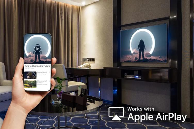 A hand holds a smart phone, showing the Apple Airplay display on it. In the background, an LG Hotel TV shows the same science fiction movie that is on the phone. In the corner of the image is the Airplay icon and text reading Works with Apple Airplay.