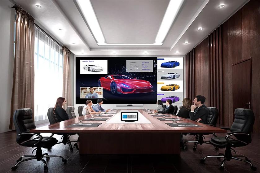 Conference room with people sitting at a table facing a large screen displaying car designs.