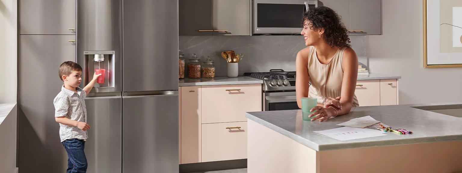 Woman standing by kitchen island and young boy dispensing water from the kitchen refrigerator dispenser.  