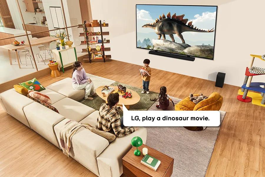 A father and three children sit around a brightly lit living room while he voice-operates the QNED99T to put a dinosour movie on the screen.