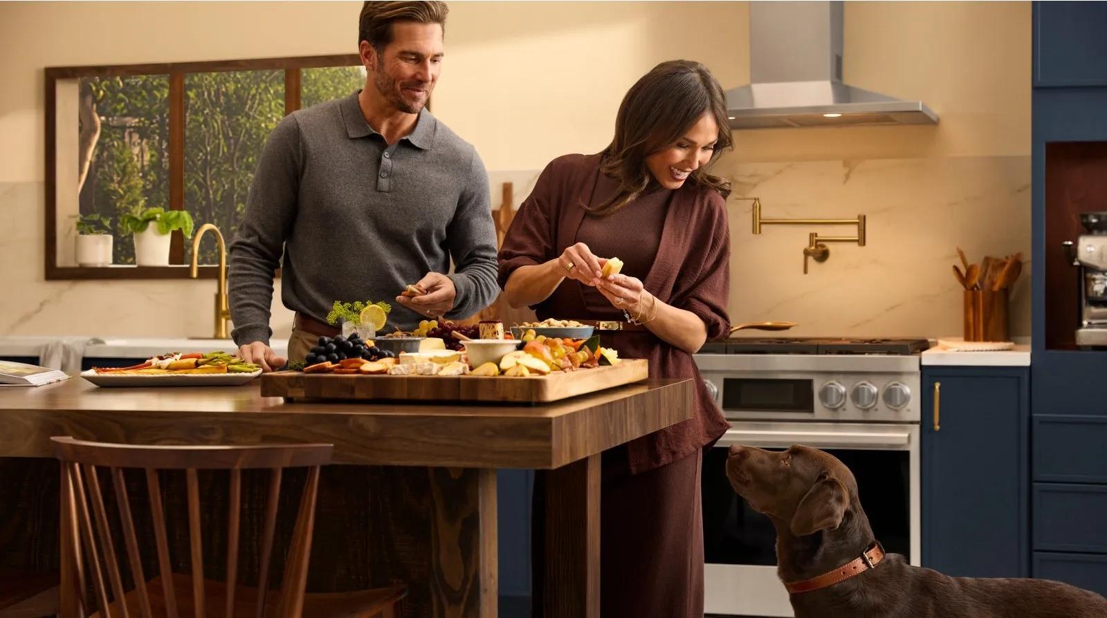 Couple preparing a charcuterie board in a navy blue kitchen with brass fixtures and a stainless steel range hood, with a dog looking on.