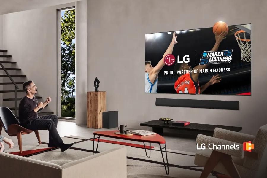 An LG TV mounted on a wall displays a close-up of a basketball player in a red jersey performing a layup being challenged by a player in a white jersey as a fan cheers on in a modern living room. Partnership lockup of LG and NCAA March Madness is overlayed on the TV. LG Channels logo.