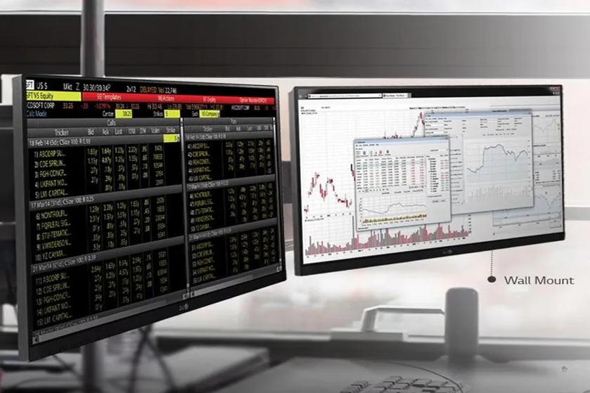 The image shows a dual-monitor setup, wall-mounted, displaying financial data and charts. The left monitor's screen is filled with rows of numbers and text, typical for a financial terminal or stock market feed. The screen uses a black background with green and white text. The right monitor displays several overlapping financial graphs and charts, with red and blue lines indicating data trends over time. The entire setup is positioned in front of a window, suggesting a modern, professional environment.