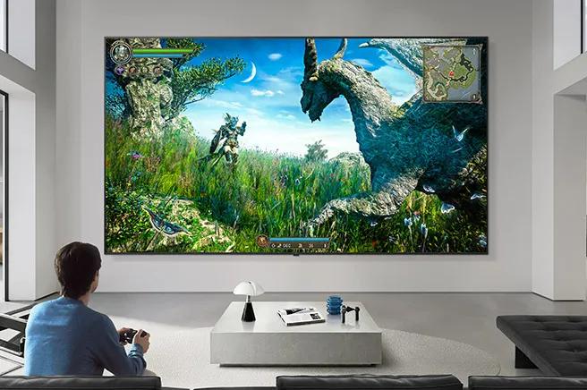 LG TV for gamers appears mounted on a wall in a black-and-white-toned living room, showing a fantasy dinosaur game scene on a large display while a person sits on a sofa holding a controller.