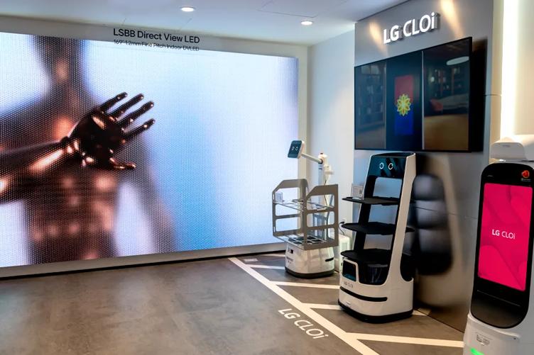 The entrance of the New Jersey Business Innovation Center. Three CLOi robots (Guidebot, ServeBot, and CarryBot) stand against the right hand wall. The wall to their right, to the left side of the image, has a large scale DVLED display mounted on it. A robotic style metallic figure is displayed in bold colors on its display.