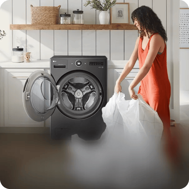 A woman in an orange dress pulling a large white comforter out of a front-loading, dark metallic LG washing machine in a bright kitchen or laundry space.