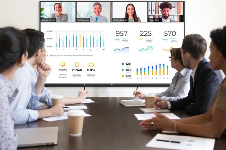 A group of professionals at a conference table, looking at an LG CreateBoard. Displayed on the CreateBoard is a presentation with various charts and graphs, and four other business people on video call. 