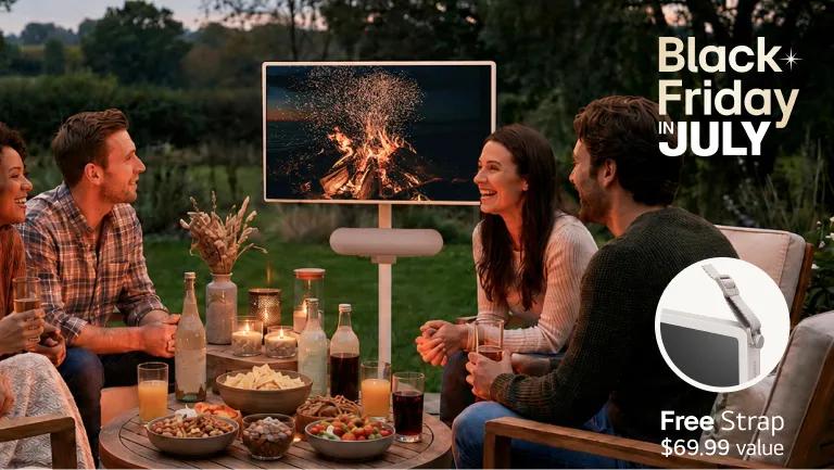 Black Friday in JULY -- LG StanbyME TV displayed on a portable stand in an outdoor patio setting with a group of friends gathered around a table with food, drinks, and candles at dusk, displaying a campfire scene. Free Strap $69.99 value.