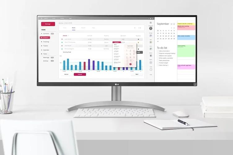 An LG Ultra Wide monitor on a clean white desk. The monitor is displaying a window with a bar chart and other information. To the right of this, another window shows a calendar, a to-do list, and a day at a glance with various meetings scheduled in colorful blocks. Near the monitor is a pen and notebook, pens in a mesh holder, some books, and a mouse and keyboard. The back of a desk chair is visible in the forefront.